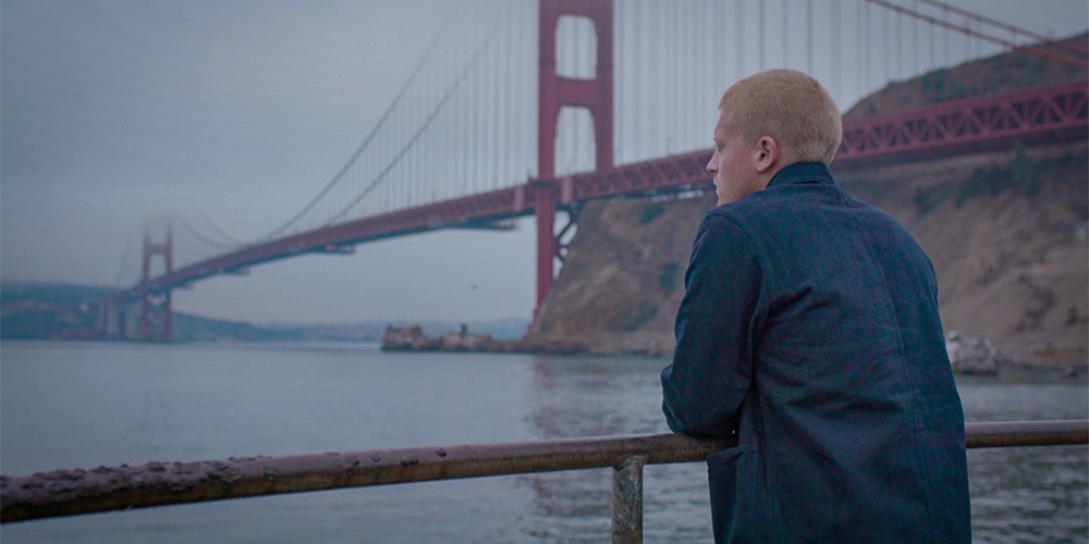 A young man with short red hair, wearing a blue jacket, in semi profile from behind with a suspension bridge in the background.