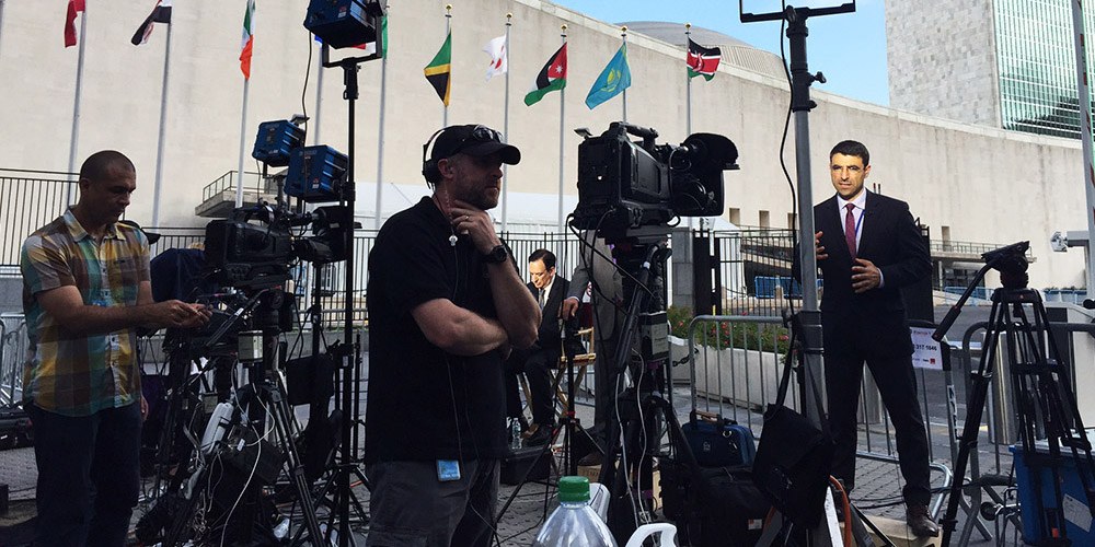 Two cameramen in foreground filming a presenter at a news event