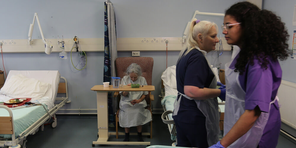 Hospital staff working on a ward while a patient sits in the background