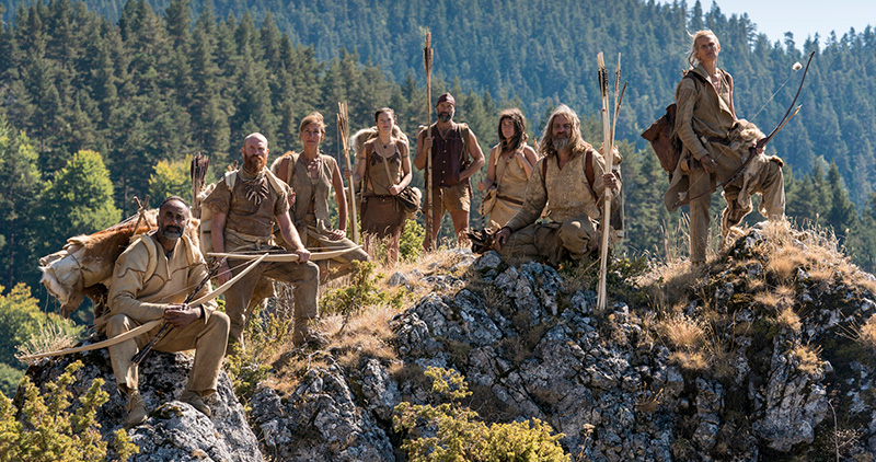 Male and female Stone Age experts, dressed as their ancestors, carrying spears and bows on a mountain top in forested wilderness